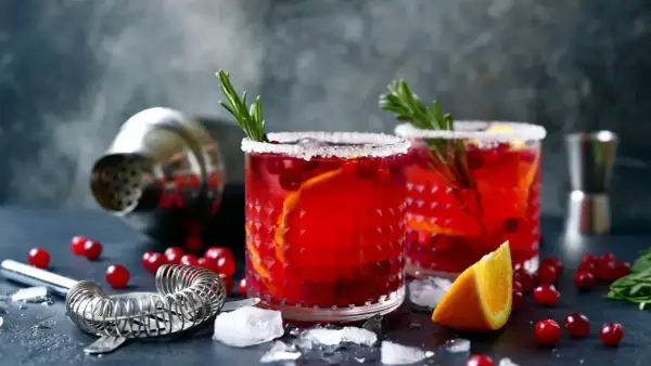 Deux verres de cocktail festif rouge vif, garni d'orange et de romarin, sur un fond sombre avec un shaker.