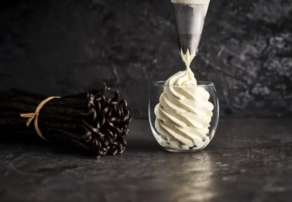 Une poche à douille dépose une crème diplomate vanillée dans un verre. Des gousses de vanille sont à côté, prêtes pour la recette.