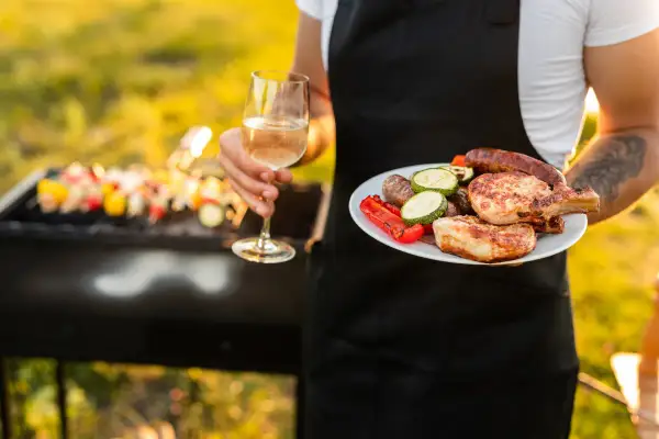 Un verre de vin blanc et une assiette de grillades, dont des côtelettes de porc et des saucisses, avec une dinde en arrière-plan sur le barbecue.