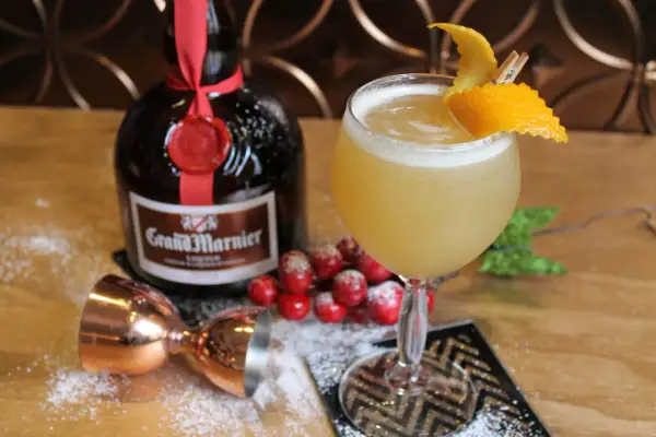 Un cocktail Grand Marnier, garni d'une écorce d'orange, à côté de la bouteille, d'un jigger en cuivre et de fausses baies rouges saupoudrées de sucre glace.